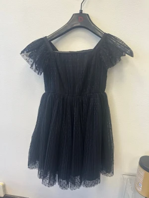 Svart kortklänning med volangärmar - Supersöt svart kortklänning med transparenta volangärmar och plisserad kjol. Klänningen har en fyrkantig urringning och är gjord i ett lätt, tunt material som ger en luftig känsla. Perfekt för dig som gillar en feminin och trendig stil.
