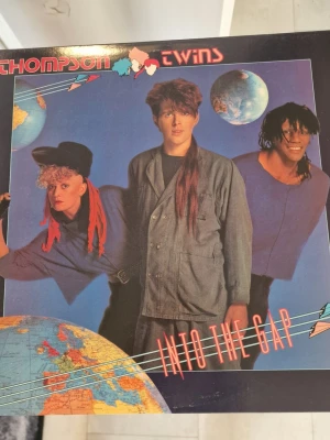 Thompson Twins Into The Gap LP - Vinylskiva med albumet 'Into The Gap' av Thompson Twins. Omslaget har en färgstark design med världskartor och bandmedlemmarna i fokus. Perfekt för dig som gillar 80-talsmusik och samlar på klassiska popalbum. Inga repor. 