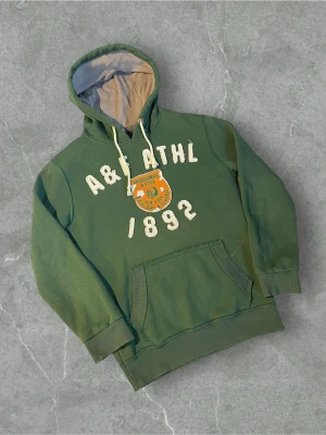 Grön hoodie från Abercrombie & Fitch | L - Snygg grön hoodie från Abercrombie & Fitch med vita bokstäver och siffror framtill samt ett broderat märke på bröstet. Liten vit fläck kolla bild 4! Det står XL men den sitter som L, skriv i privat för mått!