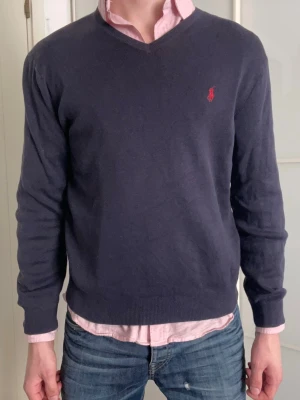 Mörkblå tröja från Polo Ralph Lauren - Klassisk mörkblå tröja från Polo Ralph Lauren i 100% pima bomull. Tröjan har V-ringning, långa ärmar och den ikoniska röda logotypen broderad på bröstet. Perfekt att bära över en skjorta för en stilren look. Storlek M men är liten i storleken