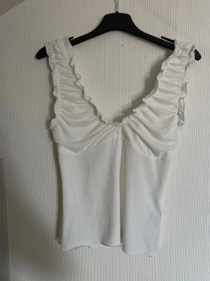 Vit topp med volangaxelband - Säljer en söt vit topp med breda volangprydda axelband och djup v-ringning. Toppen har en figurnära passform och är tillverkad i mjukt bomullsliknande material. Perfekt att styla med jeans eller kjol för en trendig look💕🥂