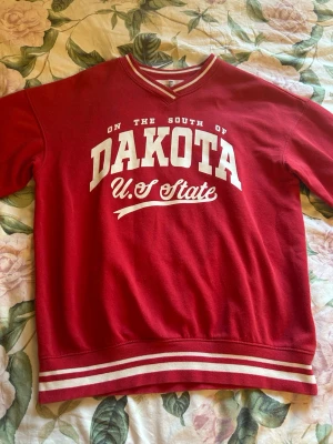 Röd college-tröja Dakota U.S State - Röd college-tröja med vit text 'DAKOTA U.S State' framtill. Tröjan har vita ränder på muddar och nederkant samt V-ringad hals med randig detalj. Långärmad och i mjukt sweatshirtmaterial, perfekt för en avslappnad stil.