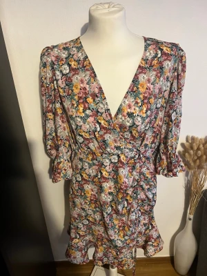 Blommig omlottklänning med volang - Söt kort blommig omlottklänning med volangkant och v-ringning. Klänningen har trekvartsärm med lätt puff och volang vid ärmslut. Färgerna går i rosa, gul, vit och grön. Perfekt för sommarens alla tillfällen!  Storlek m/l