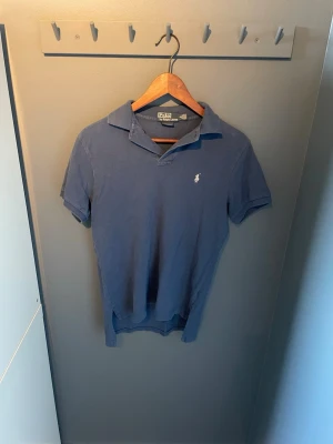 Blå pikétröja från Ralph Lauren - Klassisk blå pikétröja från Ralph Lauren med vit broderad logga på bröstet. Tröjan har korta ärmar, krage och knappar vid halsen. Tillverkad i mjuk bomull och har en custom fit passform som sitter snyggt på kroppen.