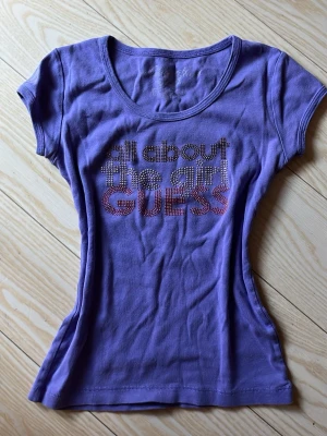 Lila t-shirt från Guess  - Snygg lila t-shirt från Guess med text i glittriga stenar på bröstet. Modellen är figurnära med rund halsringning och korta ärmar. På bilden syns färgerna inte så bra, men de är mer synbara i verkligheten. Aldrig använd men köpt second hand. Endast en sten har trallat av men annars finns det inga defekter. Står ej storlek men skulle säga xs-m (det finns mycket stretch)
