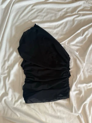 Svart oneshoulder topp i mesh - Trendig svart oneshoulder topp i meshmaterial med tight passform och rynkade detaljer. Toppen är croppad och har en asymmetrisk axel som ger en cool look. Perfekt att styla med kjol eller jeans för en snygg outfit.