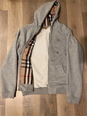 Burberry hoodie - Snygg grå zip-hoodie från Burberry med klassiskt rutigt foder i huvan och längs dragkedjan. Hoodien har dragsko i huvan, fickor framtill och ribbade muddar. Perfekt för dig som vill ha en stilren men ändå lyxig look.