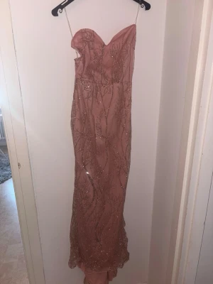 Rosa långklänning med paljetter - Elegant rosa långklänning hjärtformad urringning. Klänningen är dekorerad med glittriga paljetter i ett mönster över hela tyget och har en figurnära passform. Perfekt för bal eller festliga tillfällen.