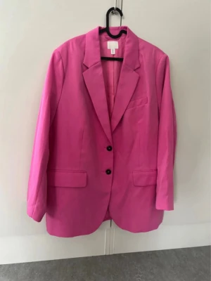 Oversized rosa kavaj från H&M - Snygg oversized kavaj i stark rosa färg från H&M. Klassisk modell med två knappar framtill, breda slag och två fickor med lock. Perfekt för att addera färg till din outfit och ge en trendig touch. Tillverkad i ett slätt tyg med mjuk känsla.