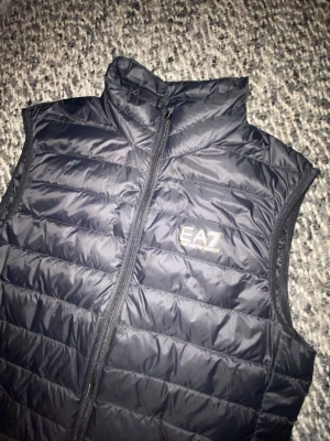 Svart dunväst från EA7 Emporio Armani - Snygg svart dunväst från EA7 Emporio Armani med guldfärgad logga på bröstet. Västen har hög krage, dragkedja framtill och quiltad design. Perfekt för lager på lager och streetwear-stil.