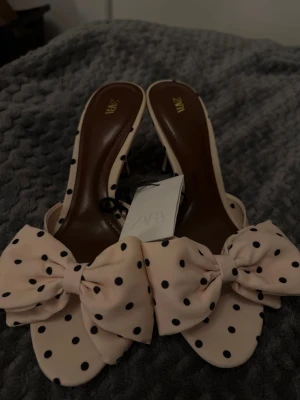 Prickiga sandaler med rosett från Zara - Säljer ett par ljusrosa sandaler från Zara med stora dekorativa rosetter framtill och svarta prickar. Sandalerna har öppen tå och låg klack, samt en mjuk innersula i brunt. Perfekta för dig som vill sticka ut med en lekfull och trendig look.