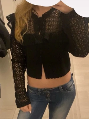 Svart spetsblus med volangdetaljer - Trendig svart blus i spets med volangdetaljer och knytning vid halsen. Blusen är genombruten med långa ärmar och har en croppad passform. Perfekt för dig som gillar en romantisk och edgy stil. Materialet känns lätt och luftigt. Storlek XL men passar mig som XS! 