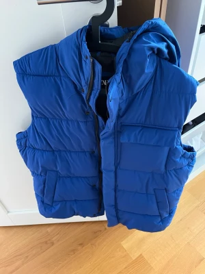 Blå dunväst med huva från Zara - Snygg blå dunväst från Zara med stora fickor framtill och huva. Västen stängs med dragkedja och tryckknappar. Mycket bra skick och ser ut som ny