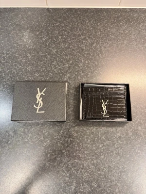  korthållare - Snygg svart korthållare glansigt läder med krokodilpräglad yta. Framsidan har det ikoniska logotypen i silver. Perfekt för kort och sedlar, levereras i originalask.