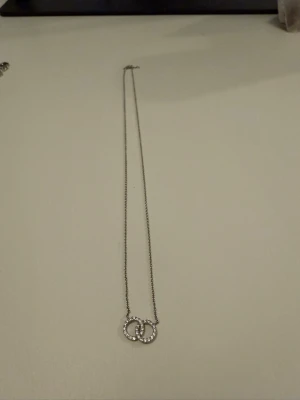 Silverfärgat halsband med pärldetaljer - Snyggt halsband i silverfärg med två sammanlänkade ringar dekorerade med små pärlliknande stenar. Kedjan är tunn och elegant, perfekt för att ge en stilren touch till din outfit.
