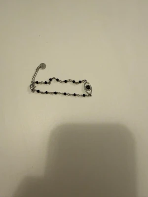 Silverfärgat armband med pärlor och öga - Snyggt armband i silverfärg med små svarta pärlor och ett dekorativt öga som detalj. Kedjan är tunn och justerbar med en liten bricka vid låset. Perfekt accessoar för dig som gillar unika smycken.
