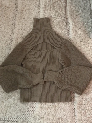 Beige ribbad stickad polotopp - Trendig beige stickad polotopp med ribbad struktur och unik öppen detalj över bröstet. Långa ärmar med breda muddar och hög polokrage ger en cool och modern look. Perfekt att styla med jeans eller kjol.