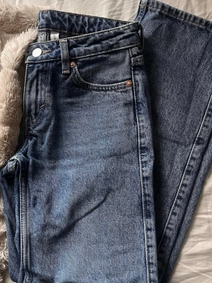 Blå raka jeans från Weekday - Säljer ett par klassiska blå jeans från Weekday med rak passform och fem fickor. Jeansen har låg midja, silverfärgade knappar och nitar samt tydliga sömmar. Tillverkade i kraftig denim som ger en tidlös look.