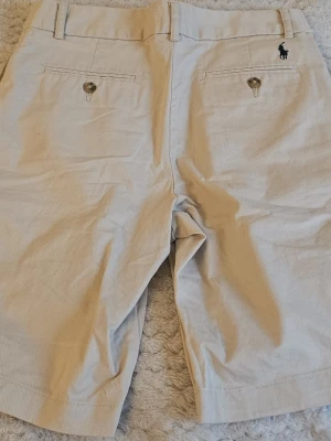Beige shorts från Polo Ralph Lauren - Klassiska beige shorts från Polo Ralph Lauren med två bakfickor med knapp och bälteshällor. Diskret broderad logga ovanför ena bakfickan. Perfekta för varma dagar och enkel att matcha med olika stilar.