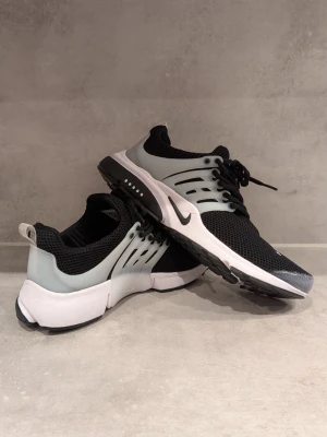 Nike svartvita mesh sneakers - Snygga sneakers från Nike med svart mesh-överdel, vita och grå detaljer samt vit sula. Skorna har en sportig look med klassisk Nike-logga på sidan och tå, samt svarta skosnören. Perfekta för dig som gillar stilrena och bekväma sneakers.