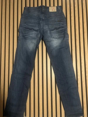 Blå jeans från Nudie Jeans - Säljer ett par klassiska blå jeans från Nudie Jeans med snyggt slitna detaljer och raka ben. Jeansen har fem fickor, kontrastsömmar och en patch med logga bak i midjan. Tillverkade i mjuk denim som sitter skönt hela dagen.