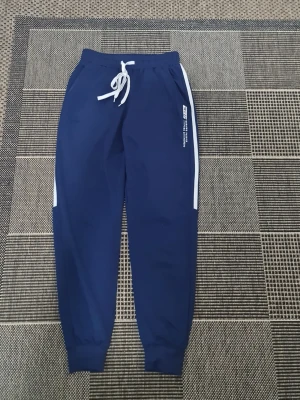 Blå mjukisbyxor med vita detaljer - Snygga blå mjukisbyxor från RED med vita sidoränder och vit dragsko i midjan. Tryck på ena benet med texten 'BURNING PASSION SURPASS DREAMS'. Perfekta för chill eller träning, med muddar vid benslut och avslappnad passform.