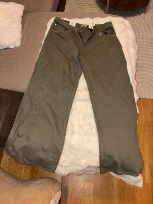 Olivgröna raka jeans - Säljer ett par olivgröna jeans med rak passform och klassisk femficksdesign. Jeansen har bälteshällor och knappgylf. Perfekta för dig som gillar en enkel och stilren look. Materialet känns slitstarkt och passar till många olika outfits.