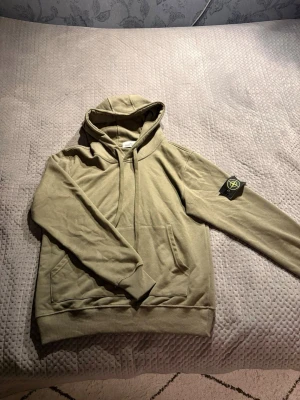 Beige hoodie från Stone Island - Säljer en beige hoodie från Stone Island med klassisk patch på vänster ärm. Hoodien har huva med dragsko, känguruficka framtill och ribbade muddar. Tillverkad i mjuk bomull för skön känsla och avslappnad look.