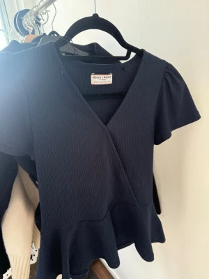 Svart topp med volang från Holly & Whyte - Trendig svart topp från Holly & Whyte by Lindex med korta puffärmar, omlott-ringning och volang nedtill. Tillverkad i ett ribbat tyg som ger en snygg struktur. Perfekt att styla med jeans eller kjol för en cool look.