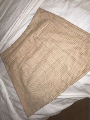 Beige rutig kortkjol med fickor - Snygg beige kortkjol med rutigt mönster och två dekorativa fickor bak. Kjolen har bälteshällor och klassisk A-linjeform. Perfekt att matcha med en enkel topp eller stickad tröja.