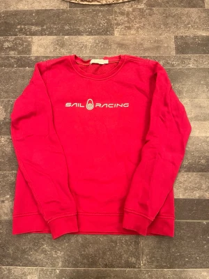 Rosa sweatshirt från Sail Racing - Rosa sweatshirt från Sail Racing med vit logotyp och text på bröstet. Tröjan har rund halsringning, långa ärmar och ribbade muddar vid ärmslut och nederkant. Perfekt för en avslappnad och sportig stil.