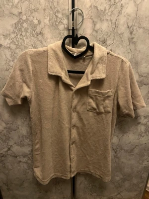 Beige kortärmad bekväm mjuk skjorta storlek S, herr och dam  - Mysig beige skjorta i mjukt frottéliknande material. Skjortan har korta ärmar, klassisk krage, knappstängning framtill och en bröstficka. Perfekt för chilliga kvällar hemma. Perfekt för vardag eller sommarkvällar 