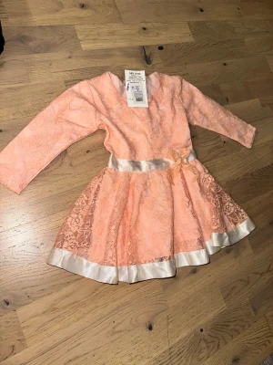 Ljusrosa spetsklänning med rosett - Söt ljusrosa klänning från S&V med spetsdetaljer, lång ärm och vacker rosett i midjan. Kjolen har volang och vita satinkanter nertill. Perfekt för dig som gillar romantisk stil och fina detaljer.