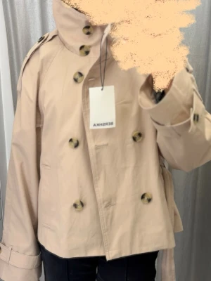 Kort beige trenchcoat från Bershka - Stilren beige trenchcoat från Bershka med dubbelknäppning, bred krage och knytskärp i midjan. Jag vill gärna byta till storlek xs eller s!