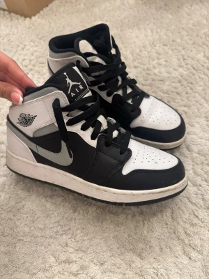 Jordan mid unisex - Säljer ett par Air Jordan 1 Mid sneakers i svart och vitt med klassisk Nike Swoosh i grått. Skorna har snörning, perforerad tåbox och ikoniska Jordan Wings-loggan på sidan. Tillverkade i läder och textil för en clean och tidlös look.