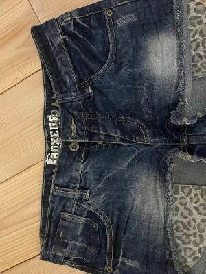 Blå jeansshorts från Boxeur Des Rues - Säljer ett par blå jeansshorts från Boxeur Des Rues med slitna detaljer och råa kanter. Shortsen har leopardmönstrat tyg på insidan av benen och klassiska fem fickor. Perfekta för en avslappnad och cool stil.