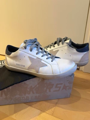 Golden Goose sneakers - Golden Goose Superstar sneakers i vitt läder med beige mockastjärna på sidan, grå skosnören och mörkblå häl.  Dustbag, GGDB-passport och låda följer med. Använda 2 ggr. Skriv vid minsta fundering!