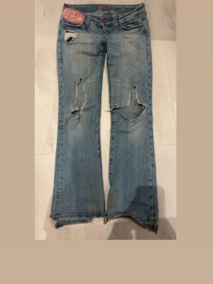 Ljusblå slitna bootcut jeans - Säljer ett par ljusblå bootcut jeans med stora slitningar på knäna och fransiga benslut. Jeansen har dekorativa bakfickor med broderi och knappdetaljer. Perfekta för en avslappnad och trendig look. Storlek 26, midjemått: 36 och Innerbenslängd: 77 