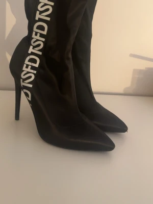 Svarta stövletter med TSFD-tryck - Snygga svarta stövletter med spetsig tå och hög smal klack. Skorna har ett vitt TSFD-tryck längs skaftet och är tillverkade i ett glansigt tygmaterial som ger en trendig look.