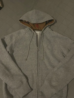 Grå zip-hoodie från Burberry - Snygg grå zip-hoodie från Burberry med klassiskt rutigt foder i huvan. Mjuk bomullsblandning, dragsko i huvan och ribbade muddar. Perfekt för en chill och stilren look. Har en bild på mig ha på den om ni vill se hur den passar mig. 