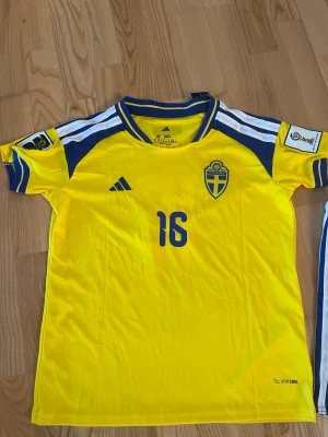 Sverige hemmaställ VM 2026 - Köpte fel storlek så behöver sälja vidare. Aldrig använd. Sveriges gula hemmaställ från Adidas med blå detaljer, nummer 16 och namnet Nygren på ryggen. Svenska landslagets emblem på bröstet och officiella VM märken på ärmarna. Tillverkad i lätt och ventilerande material med ClimaCool-teknologi. Shorts följer med!