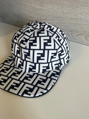 Vit och svart Fendi keps - Snygg keps från Fendi i vit och svart med det klassiska FF-mönstret över hela kepsen. Rak skärm och strukturerad form, perfekt för att lyfta din streetwear-look. Tillverkad i bomull för en bekväm känsla.