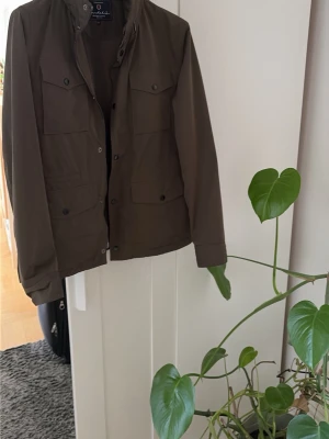 Olivgrön jacka från Bondelid - Snygg olivgrön jacka från Bondelid med fyra framfickor och hög krage. Jackan har knappar och dragkedja framtill, perfekt för dig som gillar en stilren och klassisk look. Passar till många olika outfits och är lätt att matcha.