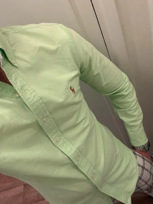 Ljusgrön skjorta från Ralph Lauren - Snygg ljusgrön skjorta från Ralph Lauren med klassisk button-down krage och broderad logga på bröstet. Skjortan har lång ärm och knäppning framtill. Perfekt för dig som vill ha en fräsch och stilren look.