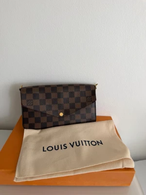Louis Vuitton Pochette Félicie - Snygg kuvertväska från Louis Vuitton i klassiskt brunt damier-mönster med guldiga detaljer och röd insida. Väskan har en elegant kedjerem och stängs med tryckknapp. Perfekt för dig som vill ha en lyxig accessoar till din outfit.