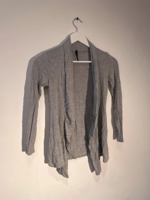 Ljusgrå tunn kofta med öppen front - Ljusgrå kofta med lång ärm och öppen front utan knappar. Tunt och mjukt material som faller snyggt längs kroppen. Perfekt att slänga över en t-shirt eller linne för en avslappnad look.