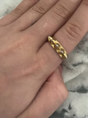 Edblad peak ring - Cool guldfärgad ring med spetsiga nitar runt hela bandet. Ringen har en unik och edgy design som sticker ut och passar perfekt för dig som gillar statement-smycken. Materialet ser ut att vara metall i guldfinish.