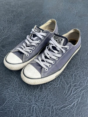 Blå Converse All Star låga sneakers - Klassiska Converse All Star sneakers i blå canvas med vita gummisulor och tåhättor. Skorna har vita snören och svart detalj runt sulan. Perfekta för en avslappnad och cool stil. Storlek 42 
