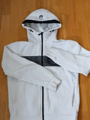 Vit hoodie från Sail Racing - Vit hoodie från Sail Racing med svart detalj över bröstet och dragkedja framtill. Hoodien har en broderad logga på huvan och praktiska fickor på sidorna. Tillverkad i mjukt material och har en sportig look med lång ärm och ribbade muddar.storlek s men passar nog även medium.