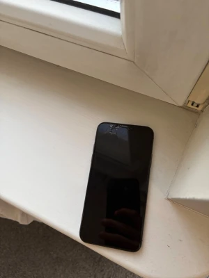 Svart iPhone X med glasbaksida - Säljer en svart iPhone X med stilren design och glasbaksida. Telefonen har en stor skärm med tunna kanter och en kamera placerad uppe till vänster på baksidan. Klassisk Apple-logga i mitten. Perfekt för dig som gillar snygg och modern teknik. 